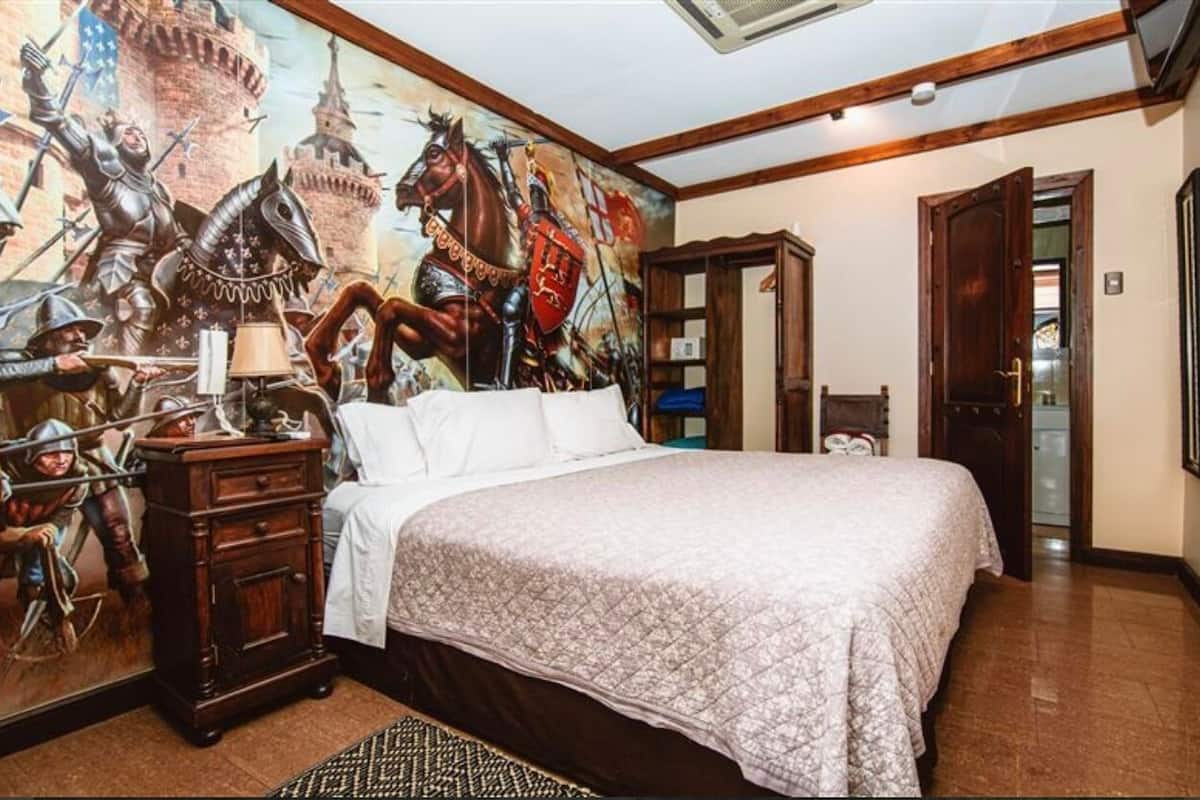 Un dormitorio con un gran mural de temática medieval, un aparador de madera, una cama con colcha estampada y una puerta que da a otra habitación.