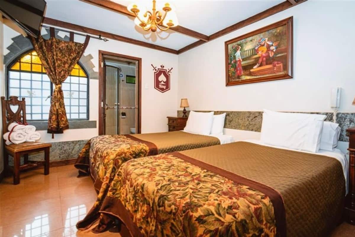 Habitación de hotel con dos camas, un candelabro, un cuadro, un perchero y una ventana con cortinas.