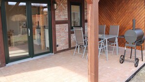 An outdoor patio with a brick floor, a glass door entrance, a brick wall, and a wood-paneled wall. There is a table with chairs and a barbecue grill. - Gemütliches Appartement in Brusow mit Garten (Kröpelin)