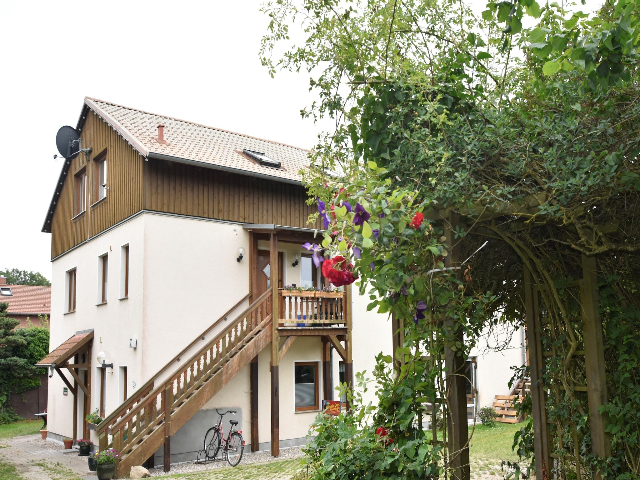 A two-story building with a wooden staircase leading to a balcony, surrounded by greenery and a garden.