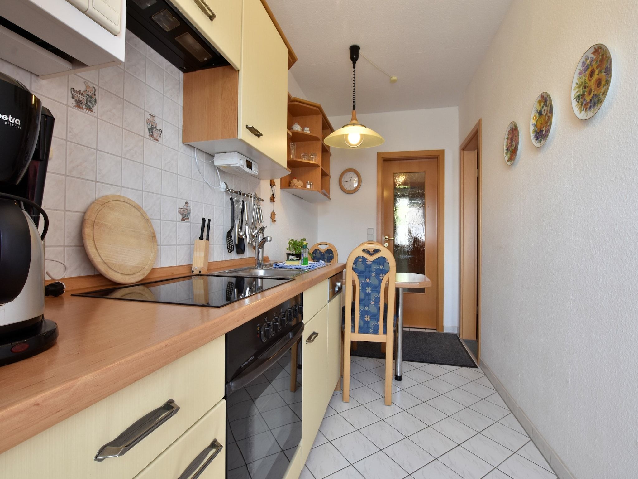 A compact kitchen with a stove, sink, and wooden counter. There is a chair and a small table with a plant.
