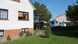 Ein weißes Gebäude mit Backsteinsockel, ein kleiner Garten mit verschiedenen Pflanzen und ein blauer Zug im Hintergrund.