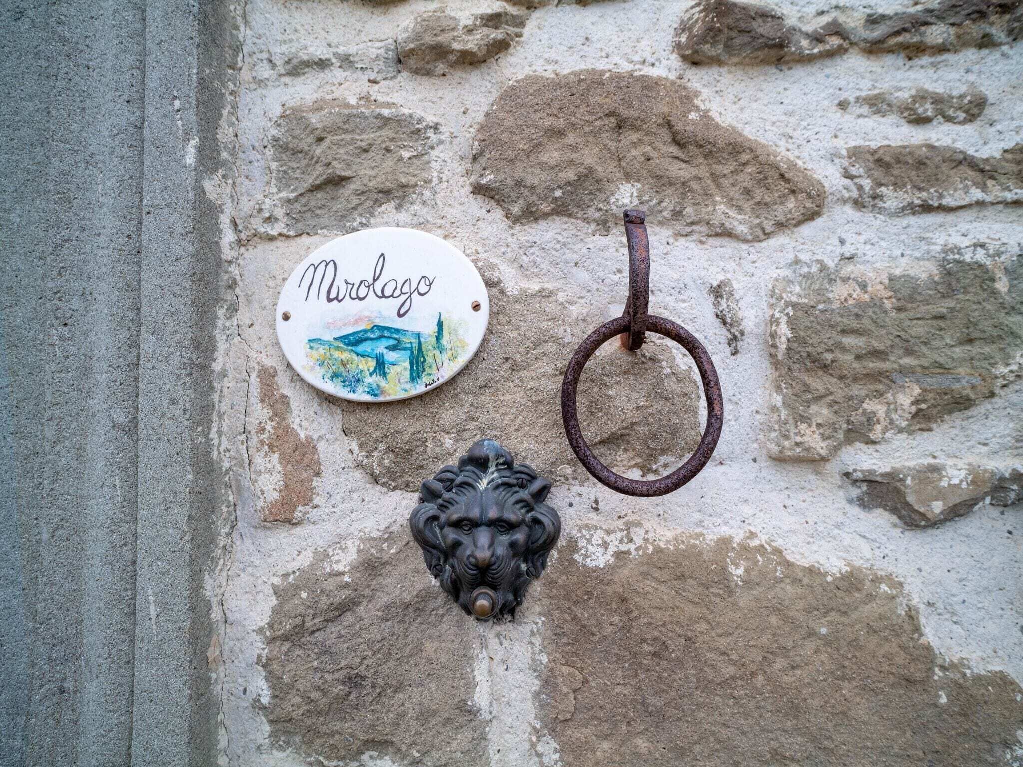 Un mur de pierre orné d’un anneau métallique, une plaque portant l’inscription « Murojago » et une peinture représentant un paysage, ainsi qu’un marteau de porte en forme de tête de lion.