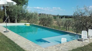 A rectangular swimming pool with stone tiles, surrounded by grass and trees, and a white umbrella.