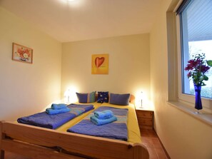 Ein Doppelbett mit blauer und gelber Bettwäsche, zwei Nachttische mit Lampen, ein herzförmiges Kunstwerk an der Wand und ein Fenster mit einem Blumenstrauß.
