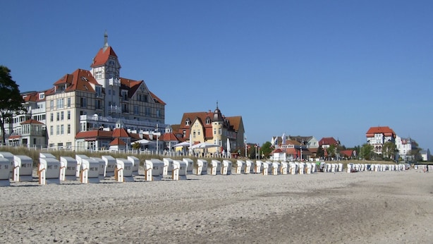 A beach with rows of beach huts labeled WE 10 to WE 17. In the background, there are buildings with red roofs and a prominent building with a tower. - Exklusiver Bungalow mit Terrasse in Rerik (Rerik)