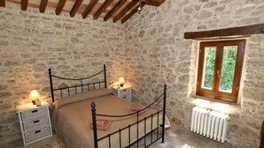 Une chambre avec des murs en pierre, un plafond en bois, un lit avec un cadre en métal, des tables de chevet avec des lampes et une fenêtre donnant sur un paysage verdoyant.