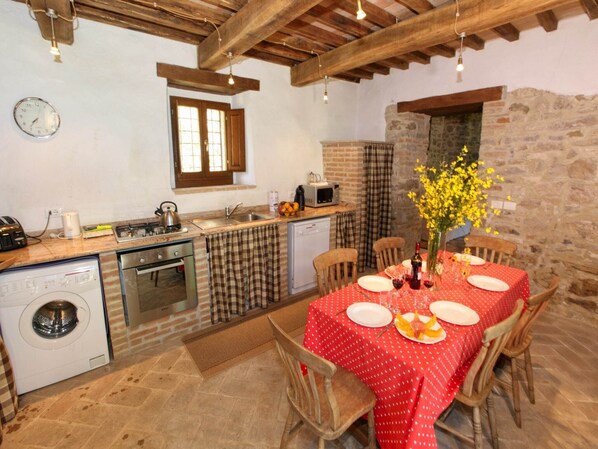 A rustic kitchen with a dining area, featuring a stone wall, wooden beams, and a red tablecloth-covered table set for two.