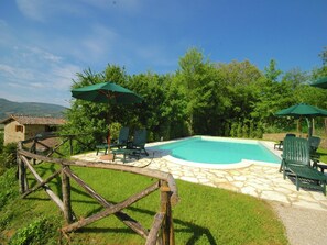 Ein Swimmingpool mit grünen Liegestühlen und Sonnenschirmen, umgeben von Steinplatten und einem Holzzaun.