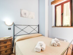 A neatly made bed with rolled towels, a wooden nightstand with a book, a wall-mounted light, and a window with curtains. - Farmhouse in Apecchio with Pool (Apecchio)
