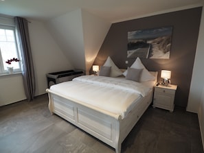 Quarto com uma cama grande, duas mesas de cabeceira, um criado-mudo, janela com cortinas e um quadro na parede.