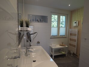 A modern bathroom with a large sink, a hairdryer mounted on the wall, a small shelf with towels, and a window with blinds. - Ferienhaus in Rerik mit Garten (Rerik)