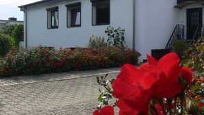 Ein weißes Gebäude mit drei Fenstern und einem schwarzen Dach, umgeben von einem Garten mit roten Blumen und Pflanzen.