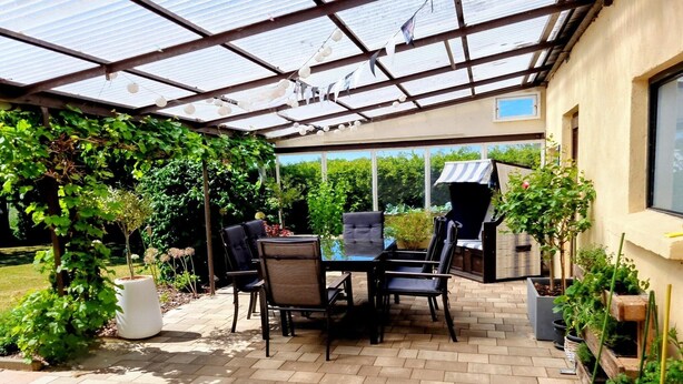 Ein überdachter Patio-Bereich mit Tisch und Stühlen, umgeben von Topfpflanzen und einem Grill.