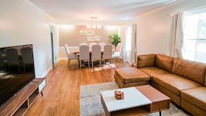 A modern living room with a wooden floor, a leather sofa, a TV, a dining table with chairs, and a chandelier. - Spacious 6 bedroom home near Downtown Macon! (Macon)