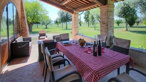 An outdoor patio with a checkered tablecloth, wicker chairs, and a view of a grassy landscape with trees.