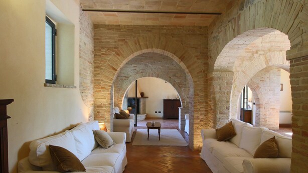 A spacious living area with two white sofas, a coffee table, and a fireplace. The room features exposed brick walls and an arched ceiling.