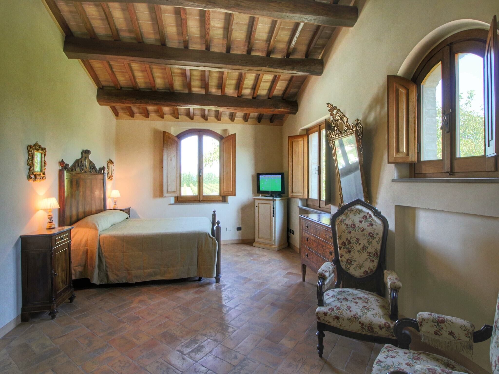 Ein Schlafzimmer mit einem Bett, Holzschrank, Kommode und einem Sessel. Ein Fernseher steht auf einem kleinen Tisch und ein Fenster mit Fensterläden ist vorhanden.