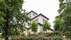 Un bâtiment à la façade symétrique, doté d’un mur de soutènement en pierre et d’un jardin où poussent différentes espèces de plantes à fleurs.