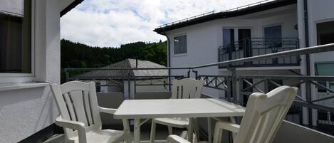 Un balcon avec une table blanche et quatre chaises, donnant sur un immeuble doté d’un balcon et offrant une vue sur une zone boisée.