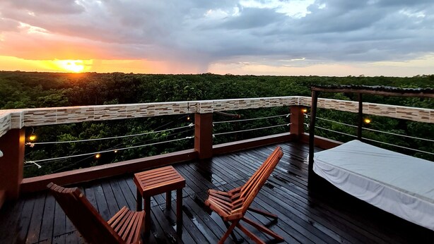 Une terrasse en bois avec une table et deux chaises, offrant une vue sur un paysage luxuriant et un coucher de soleil.