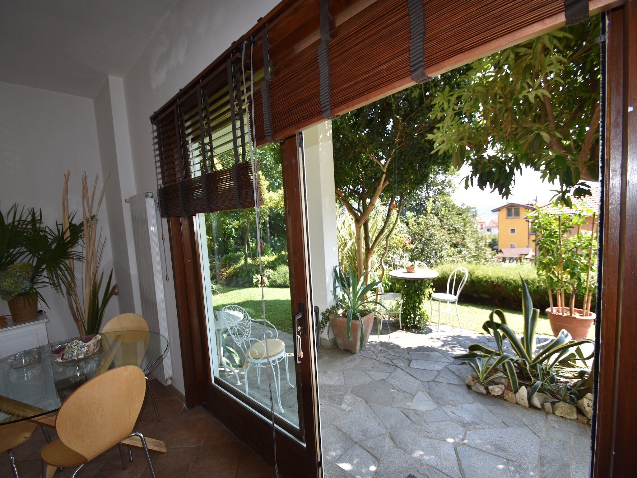 Eine Terrasse mit Glastür zu einem Garten, ein runder Tisch mit einem Stuhl und Kübelpflanzen.