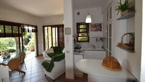 A kitchen with a gas stove, sink, and a hanging light fixture. There is a window with blinds, a chair, and a table with a green cloth.