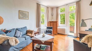 A cozy living room with a blue sofa, a wooden coffee table, and a view of greenery through large windows.