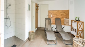 A modern bathroom with a shower, two chairs, a wooden wall, and a shelf with towels.