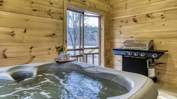 A wooden cabin interior with a hot tub, a grill, and a window with a view of trees. - In-Town Smoky Mountain Views • No Steep Roads • Fully fenced Area for Pets. (Sevierville)