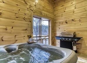 Interior de una cabaña de madera con un jacuzzi, una parrilla y una ventana con vista a árboles.