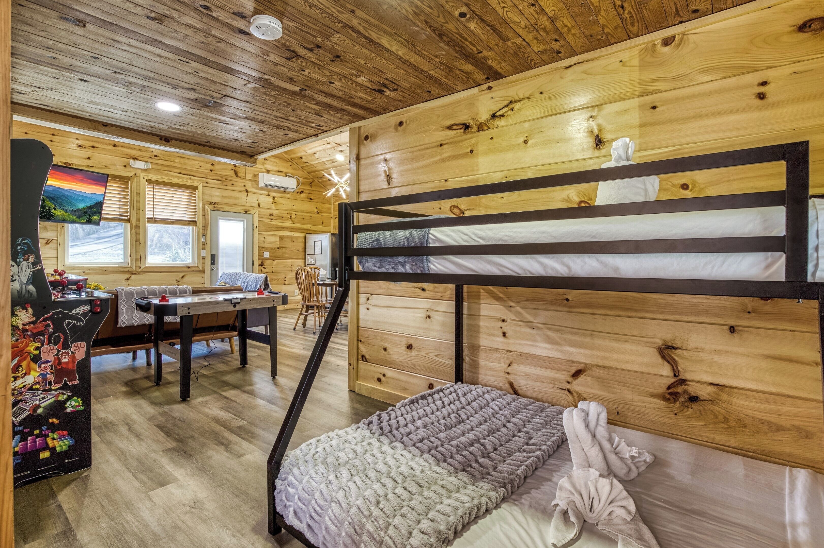A bunk bed with a wooden headboard, a desk with a chair, a table, and a window with blinds.