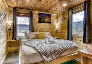 A wooden cabin bedroom with a bed, a painting, and a window with a view.