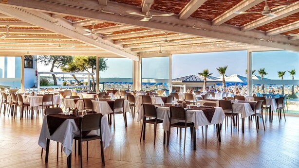 An indoor dining area with tables and chairs, large windows, and a view of a beach. - Apartment in Albarella near Beach & Activities (Albarella)