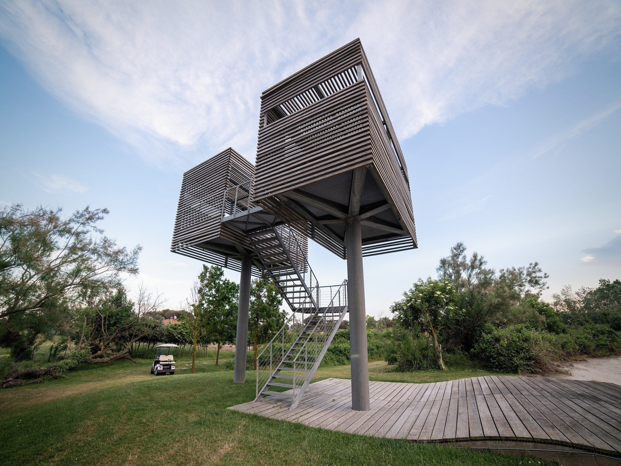 A modern outdoor structure with a wooden deck and metal framework, surrounded by greenery.