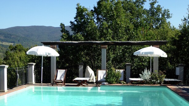A pool with lounge chairs and umbrellas, surrounded by greenery and mountains.