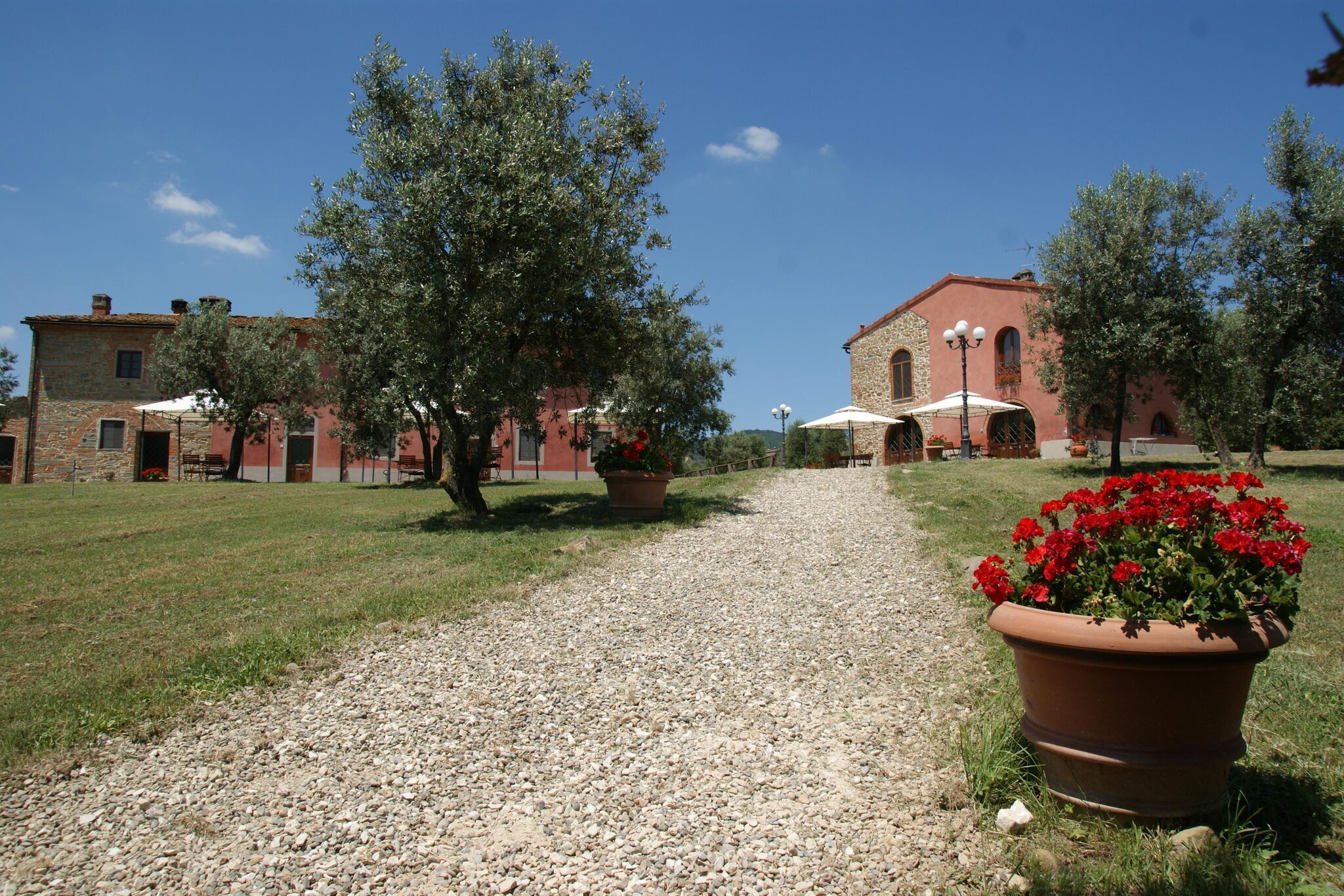Un vialetto sterrato conduce a un edificio con tetto in tegole rosse e pareti in pietra. Lungo il sentiero, vasi con fiori rossi e ombrelloni.