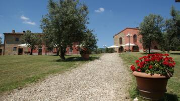 Un vialetto sterrato conduce a un edificio con tetto in tegole rosse e pareti in pietra. Lungo il sentiero, vasi con fiori rossi e ombrelloni.