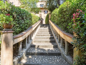 Un escalier en pierre orné d’une balustrade, entouré de verdure, avec un bâtiment en arrière-plan.