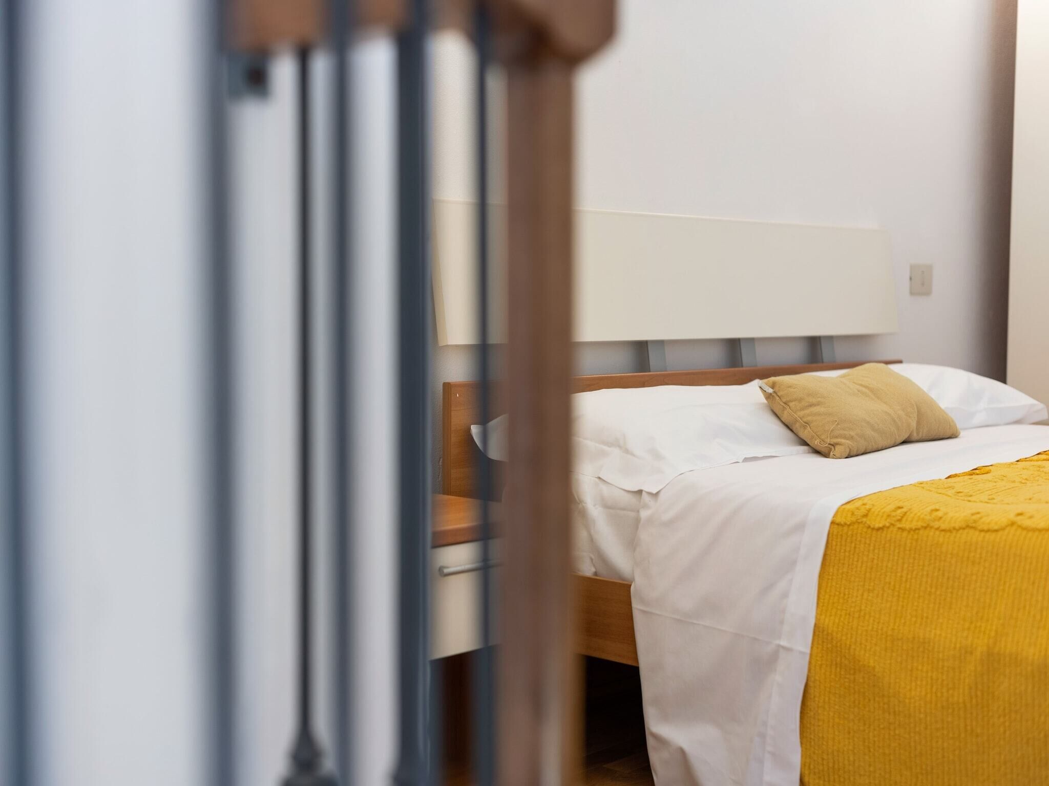 Ein ordentlich bezogenes Bett mit weißer Bettwäsche und einer gelben Tagesdecke, ein Holz-Kopfteil und eine weiße Wand.