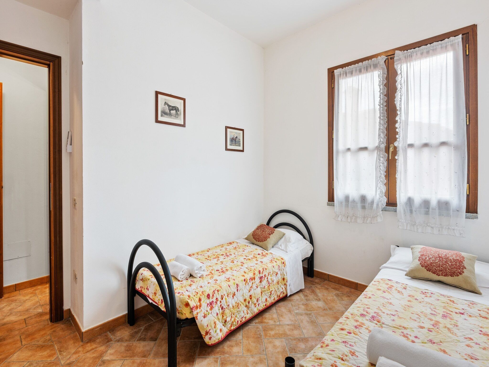 Une petite chambre bien rangée, avec un lit simple, un deuxième lit, une fenêtre avec des rideaux et deux tableaux encadrés au mur.