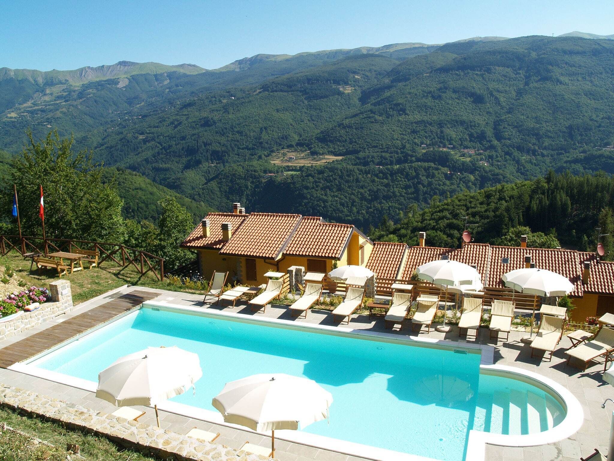 A pool area with lounge chairs and umbrellas, surrounded by buildings with red-tiled roofs, set against a backdrop of green hills.