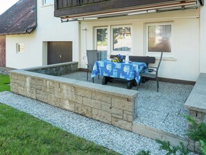 Une terrasse avec une table et des chaises, un petit jardin et un bâtiment doté d’un toit sombre et de murs blancs.