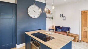 Ett modernt kök med en inbyggd köksö, en väggklocka, hängande lampor och en mysig sittgrupp med en blå soffa och kuddar.