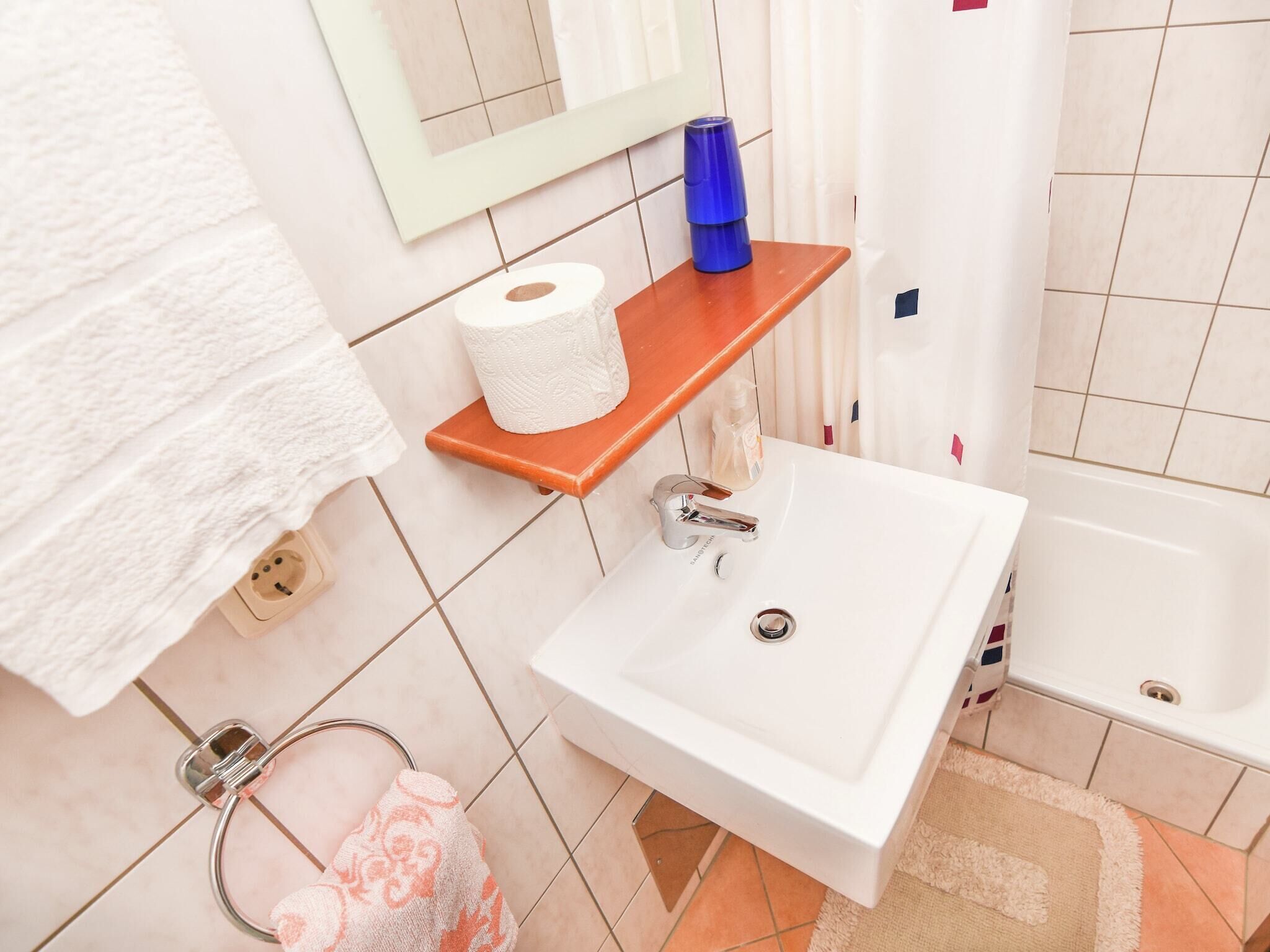 A bathroom with a white sink, a wooden shelf with a roll of toilet paper, a blue glass, and a shower curtain.