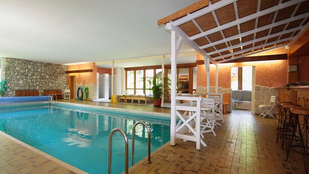 Piscina interna com parede de pedra, deck de madeira e área coberta com mesas e cadeiras.