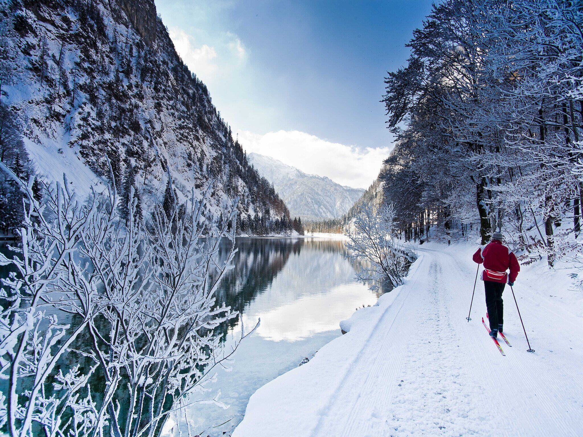 A snowy landscape with a person skiing along a snow-covered path beside a calm lake, surrounded by snow-laden trees and mountains.