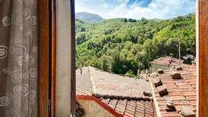 Blick aus einem Fenster mit einem gemusterten Vorhang auf eine gebirgige Landschaft mit dichten grünen Wäldern und blauem Himmel.