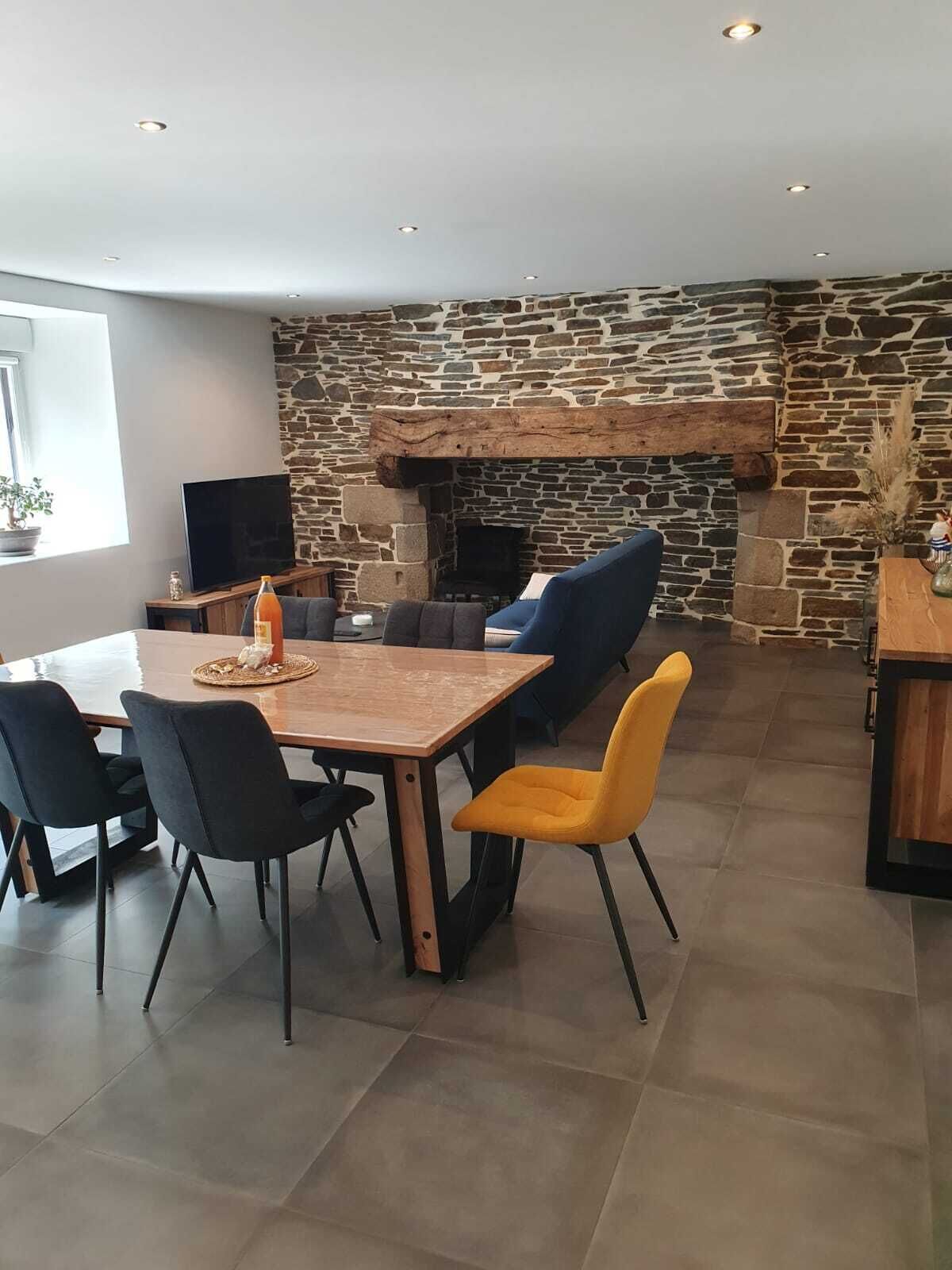 Uma sala de jantar com parede de pedra e lareira. Há uma mesa com cadeiras, uma televisão e uma garrafa de vinho.