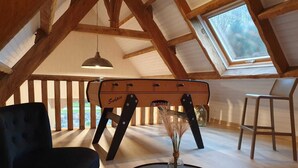 Een loft-achtige kamer met een hoge plafond, houten balken, een vintage koffer, een zwarte stoel, een ronde tafel met een vaas, en een raam met uitzicht.